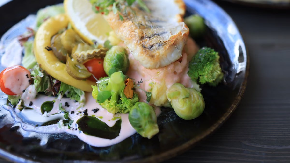 Close-up of a gourmet fish dish with assorted vegetables on a dark plate.